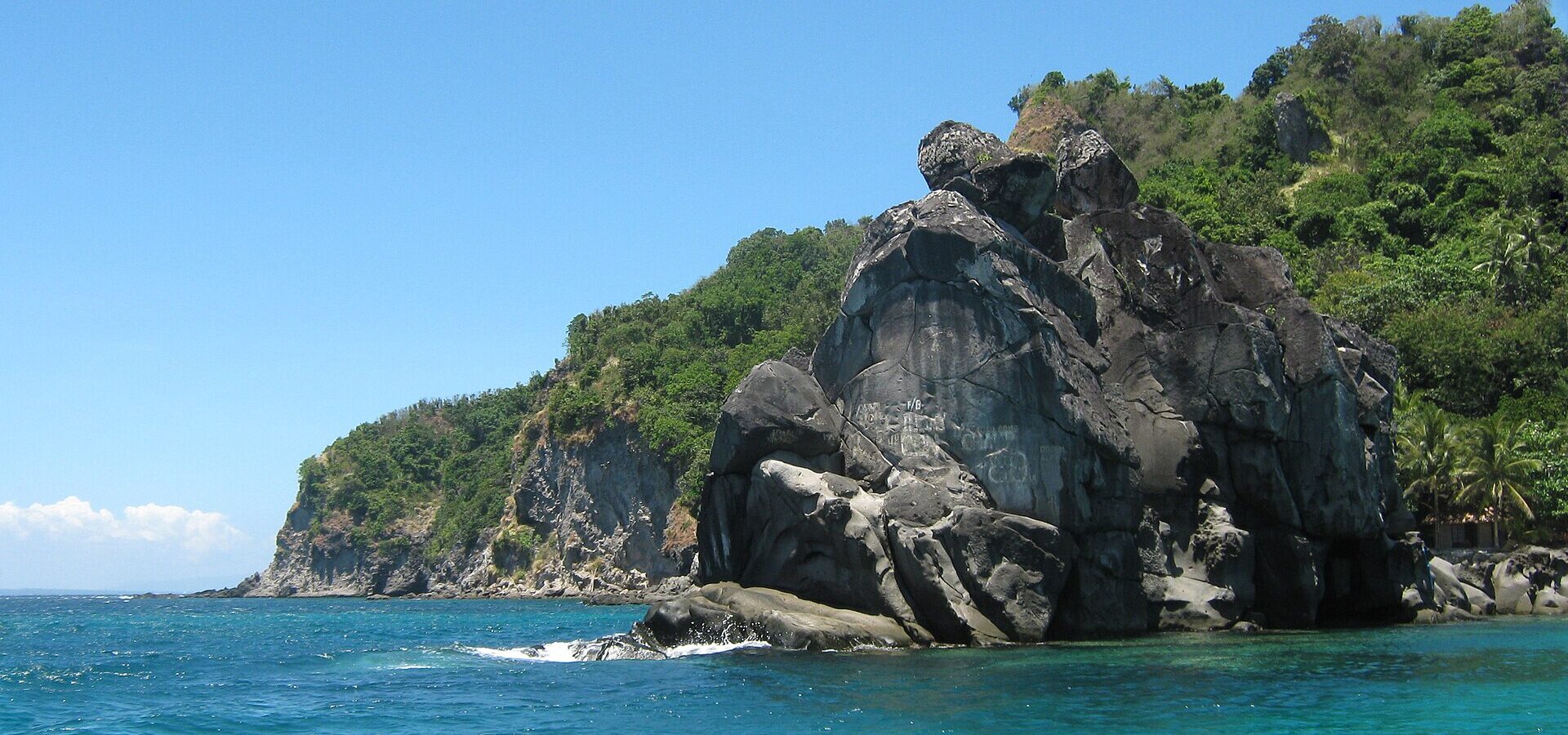 Dark volcanic rock formations and green hills above turquoise water near the boat landing area on the west side of Apo Island