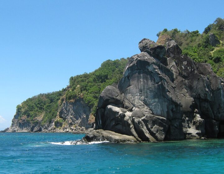 Dark volcanic rock formations and green hills above turquoise water near the boat landing area on the west side of Apo Island
