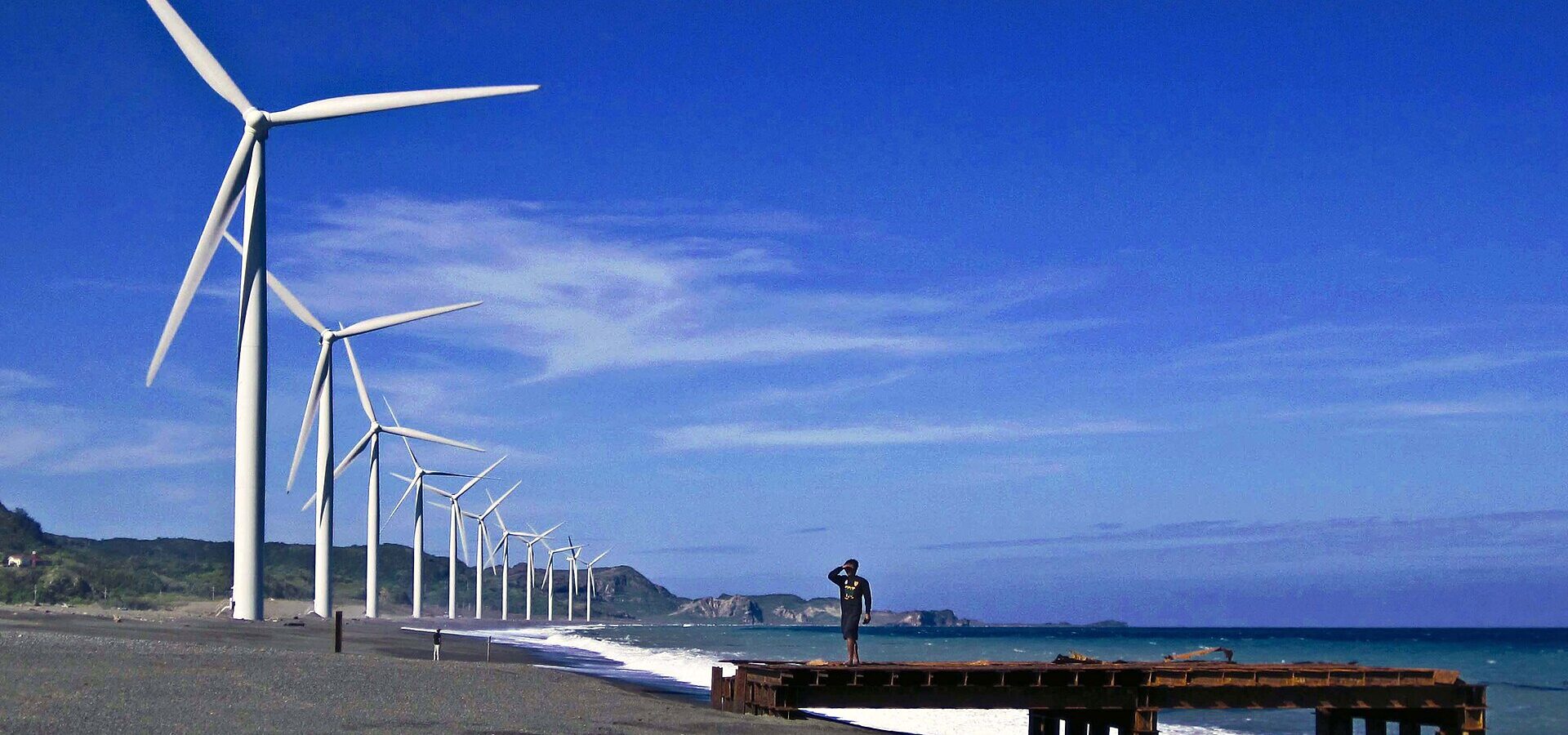 The Bangui wind turbines lined up along the grey-sand beach under a deep blue sky, with surf and a man on an old pier