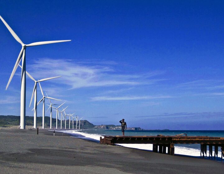 The Bangui wind turbines lined up along the grey-sand beach under a deep blue sky, with surf and a man on an old pier