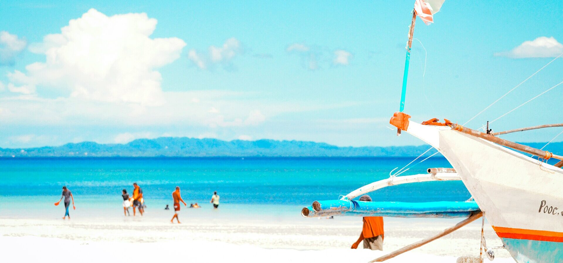 A white outrigger boat on the white sand of Bantayan Island, with people wading in shallow turquoise water
