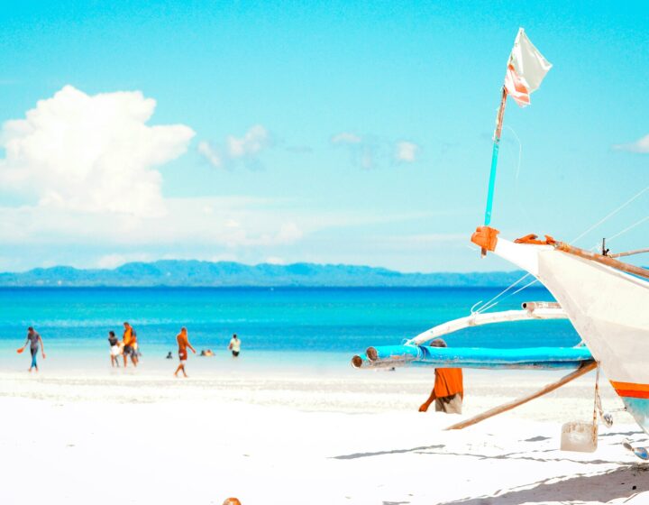 A white outrigger boat on the white sand of Bantayan Island, with people wading in shallow turquoise water