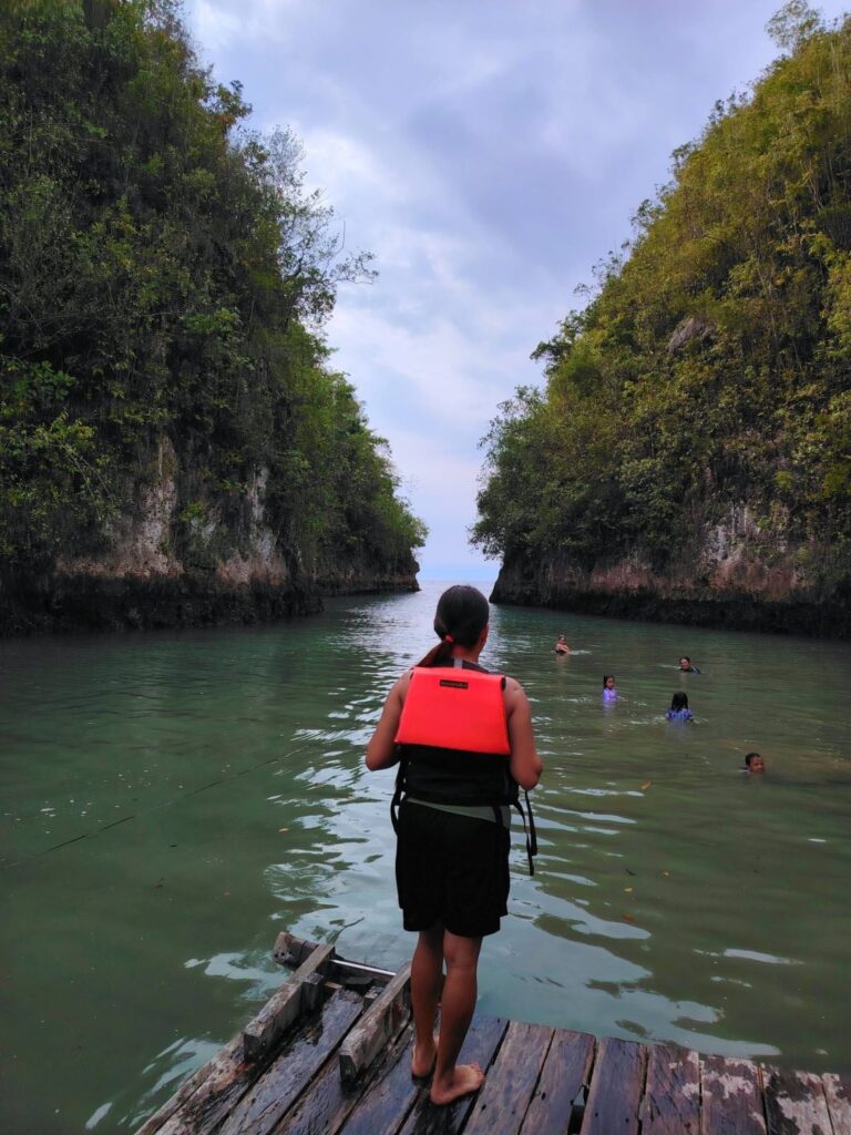 A visitor in a life vest on a wooden jetty facing the cliffs and swimmers