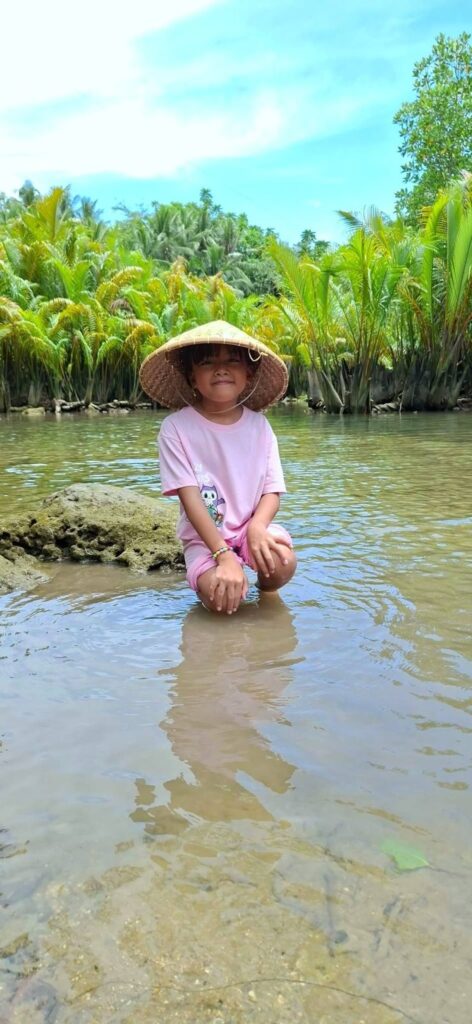 A child in a woven hat sitting in clear, shallow river water