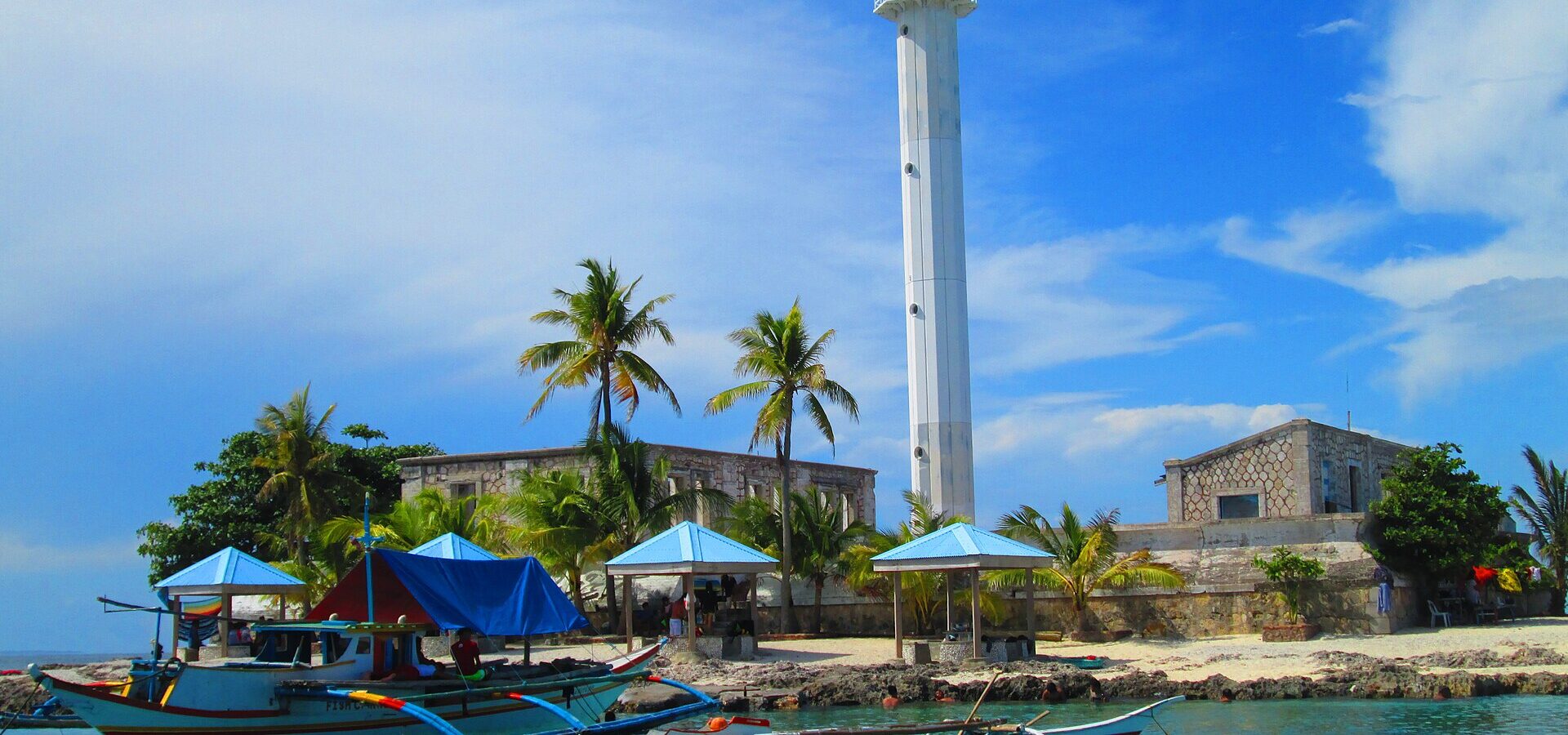 The white steel lighthouse tower on Capitancillo Island, with palms, blue huts and outrigger boats in clear turquoise water