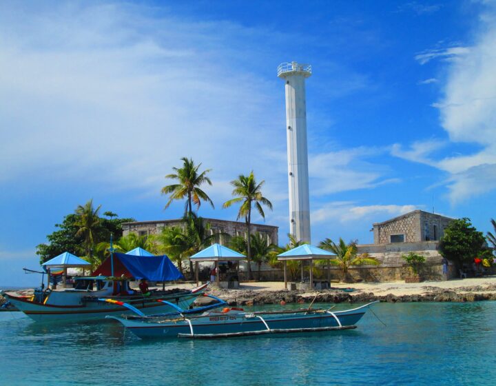 The white steel lighthouse tower on Capitancillo Island, with palms, blue huts and outrigger boats in clear turquoise water