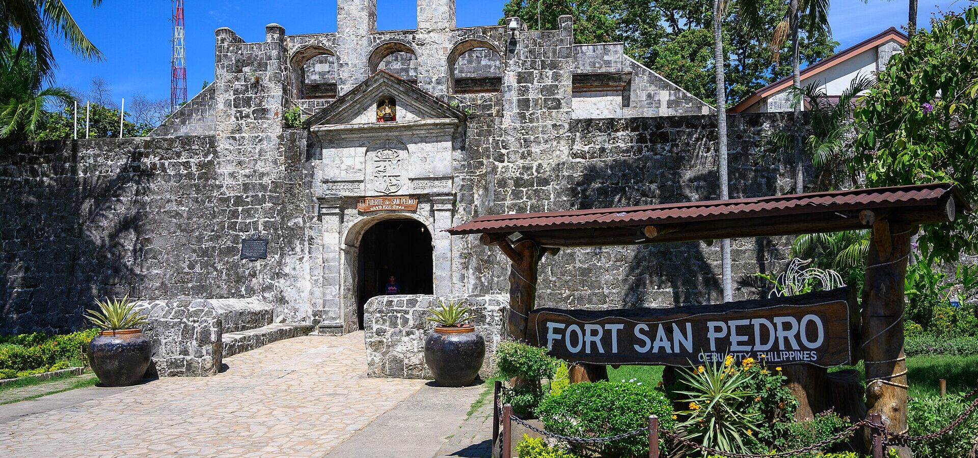 The stone main gate of Fort San Pedro under a blue sky, with the wooden FORT SAN PEDRO sign and jars of plants in front