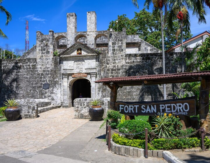 The stone main gate of Fort San Pedro under a blue sky, with the wooden FORT SAN PEDRO sign and jars of plants in front
