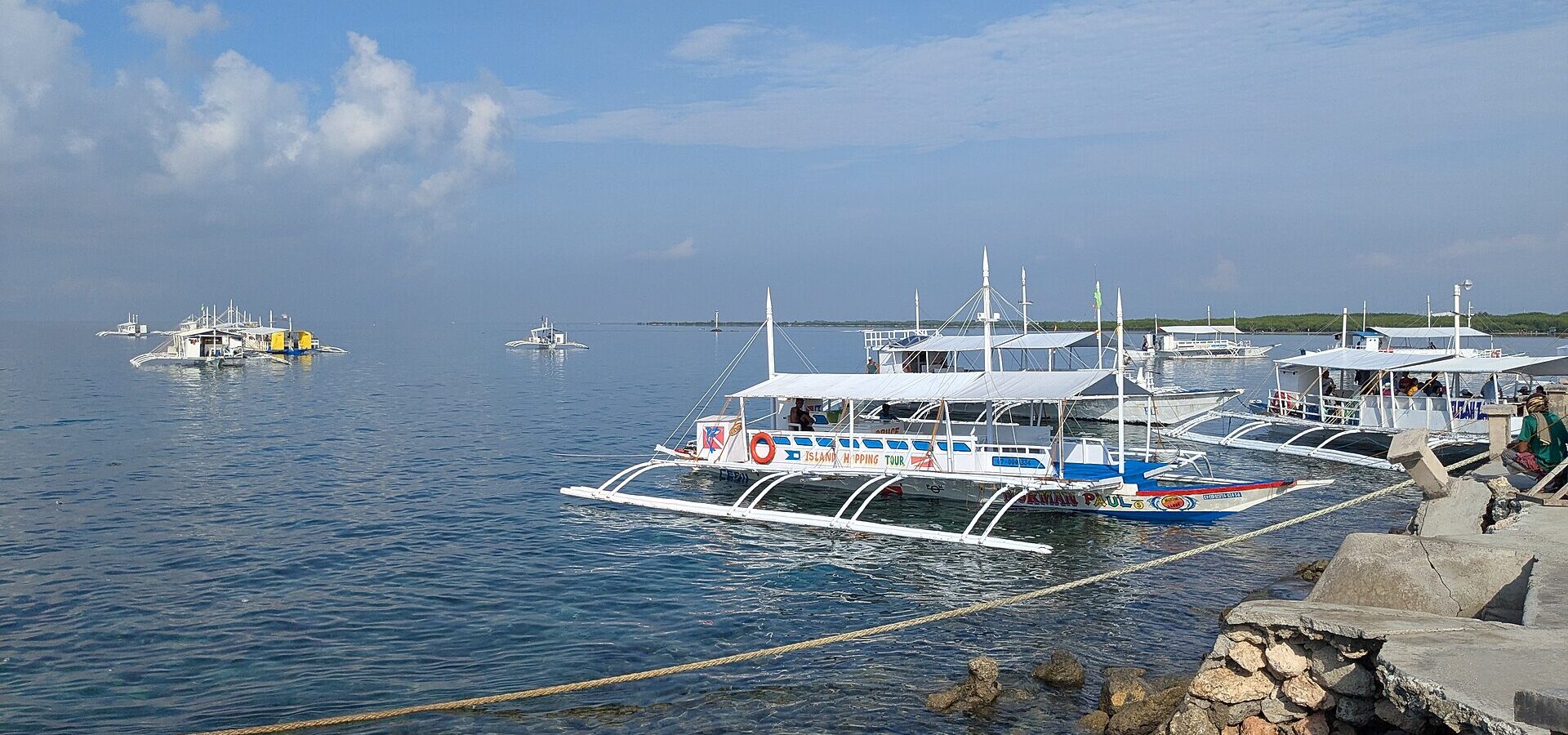 Outrigger tour boats (bangkas) moored at Cordova port, the main departure point for the Hilutungan sanctuary, under a blue sky. Shows the departure port, not the island itself