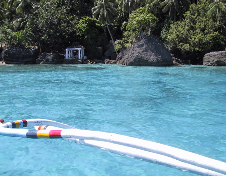 Turquoise water off the rocky, palm-covered shore of Limasawa Island, with a small white gazebo, seen from the bow of an outrigger boat