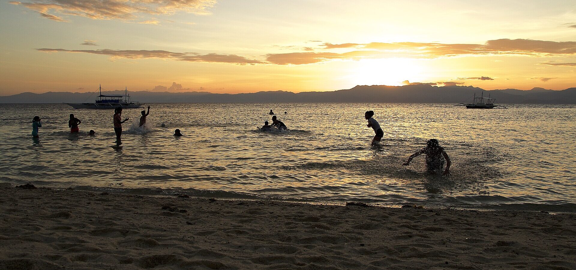 Silhouetted swimmers and bangka boats at golden sunset on Panagsama Beach, with the hills of Negros behind (5364x3016; in Commons Category:Panagsama Beach)