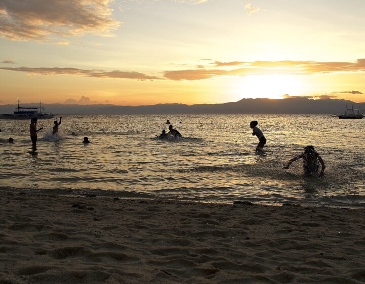 Silhouetted swimmers and bangka boats at golden sunset on Panagsama Beach, with the hills of Negros behind (5364x3016; in Commons Category:Panagsama Beach)