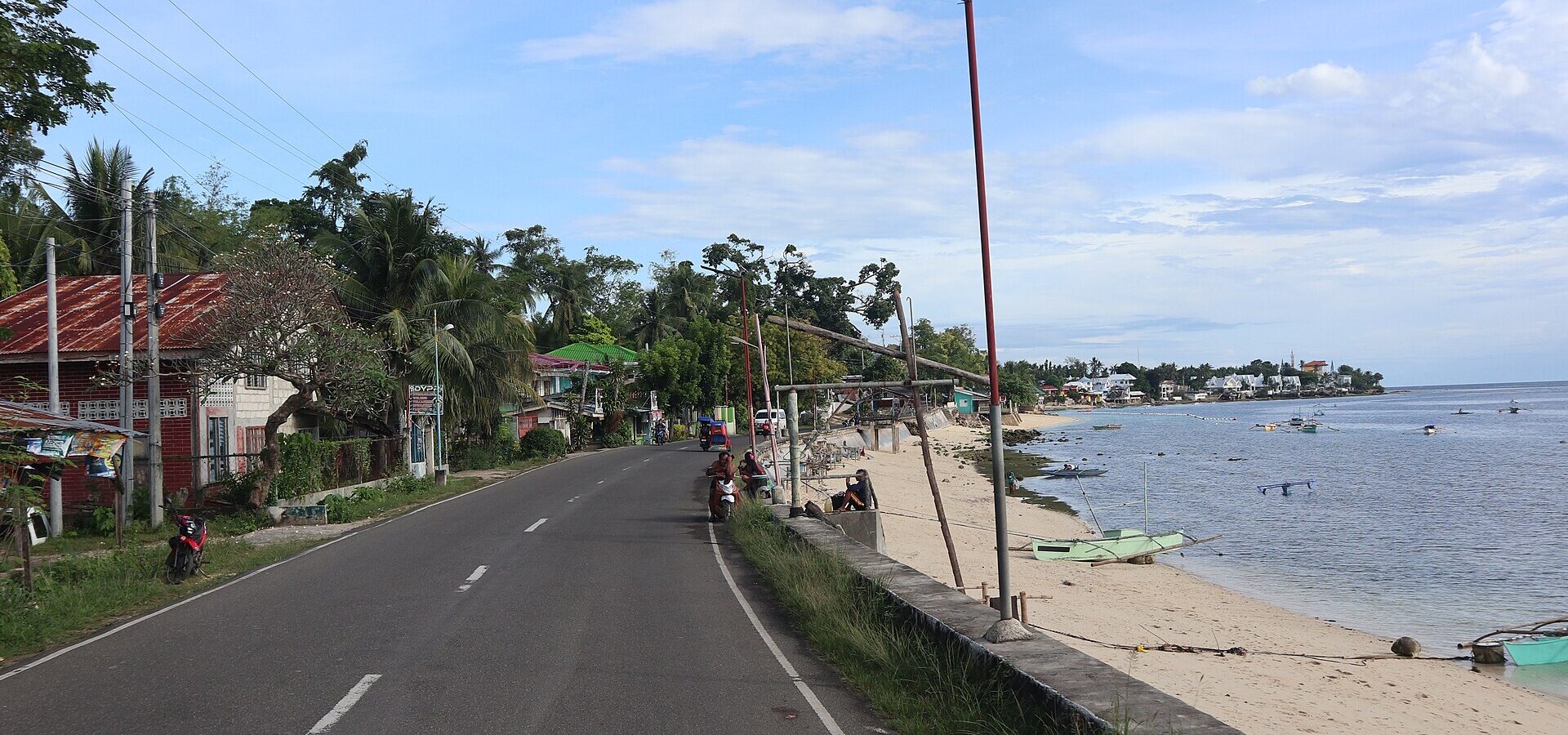 The white sand and calm blue water of Tingko Beach beside the coastal highway in Alcoy, with boats and palms