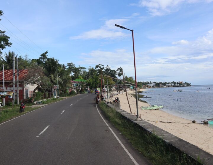 The white sand and calm blue water of Tingko Beach beside the coastal highway in Alcoy, with boats and palms