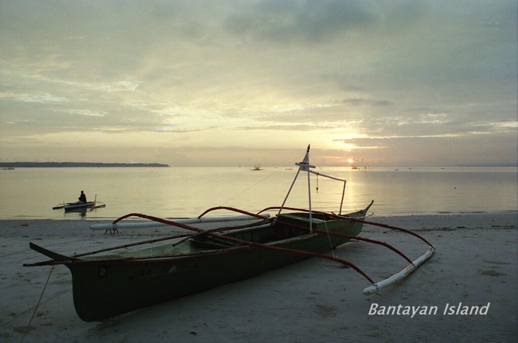 A bangka boat resting on the sand at Bantayan Island at dusk with a small boat on the calm sea beyond