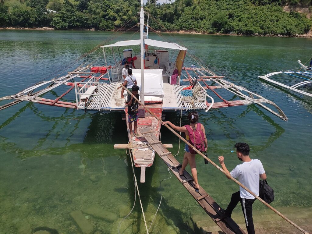 Passengers walking up a wooden plank onto an outrigger bangka boat in calm green water at Malapascua