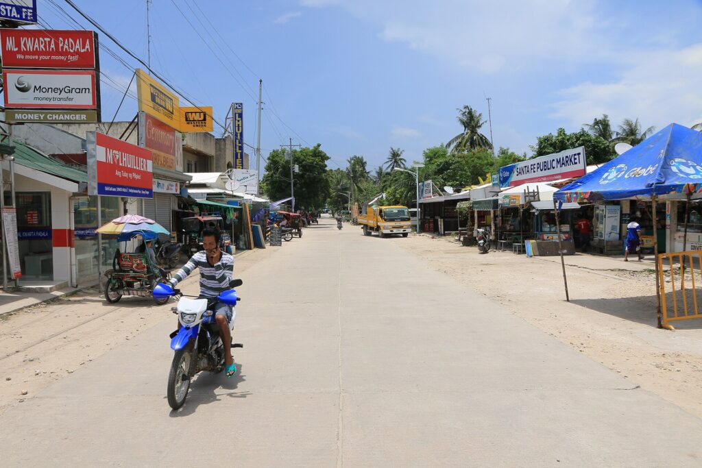 A wide sandy street in Santa Fe, Bantayan, with shops, money changers and a motorbike rider