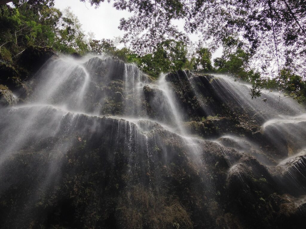 A tall curtain of water falling over a dark rock face at Tumalog Falls in Oslob, seen from below through trees